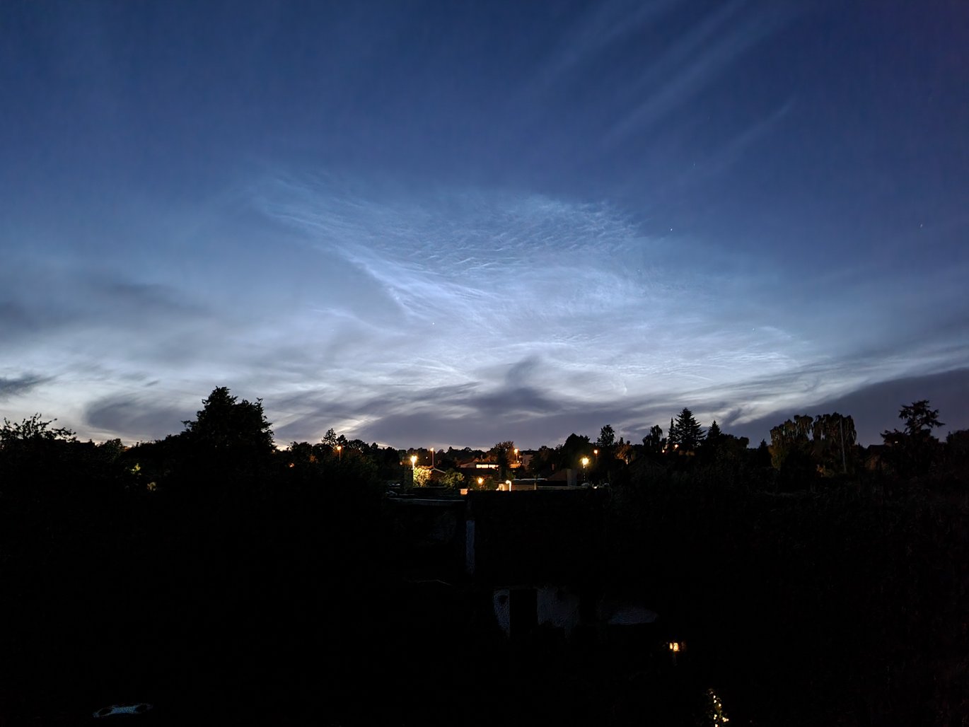Billede af lysende natteskyer set fra Åbyhøj natten til Skt. Hansaftensdag 2022. Og lave skyer midt i billedet, som er mørke, fordi de ligger nede i Jordens i skygge. Bemærk også, at der er et par stjerner, som lyser gennem skyerne. Foto: Hans Buhl.